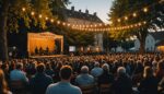 Scène de stand-up en plein air lors d'un festival d'humour régional, public assis sous des guirlandes lumineuses